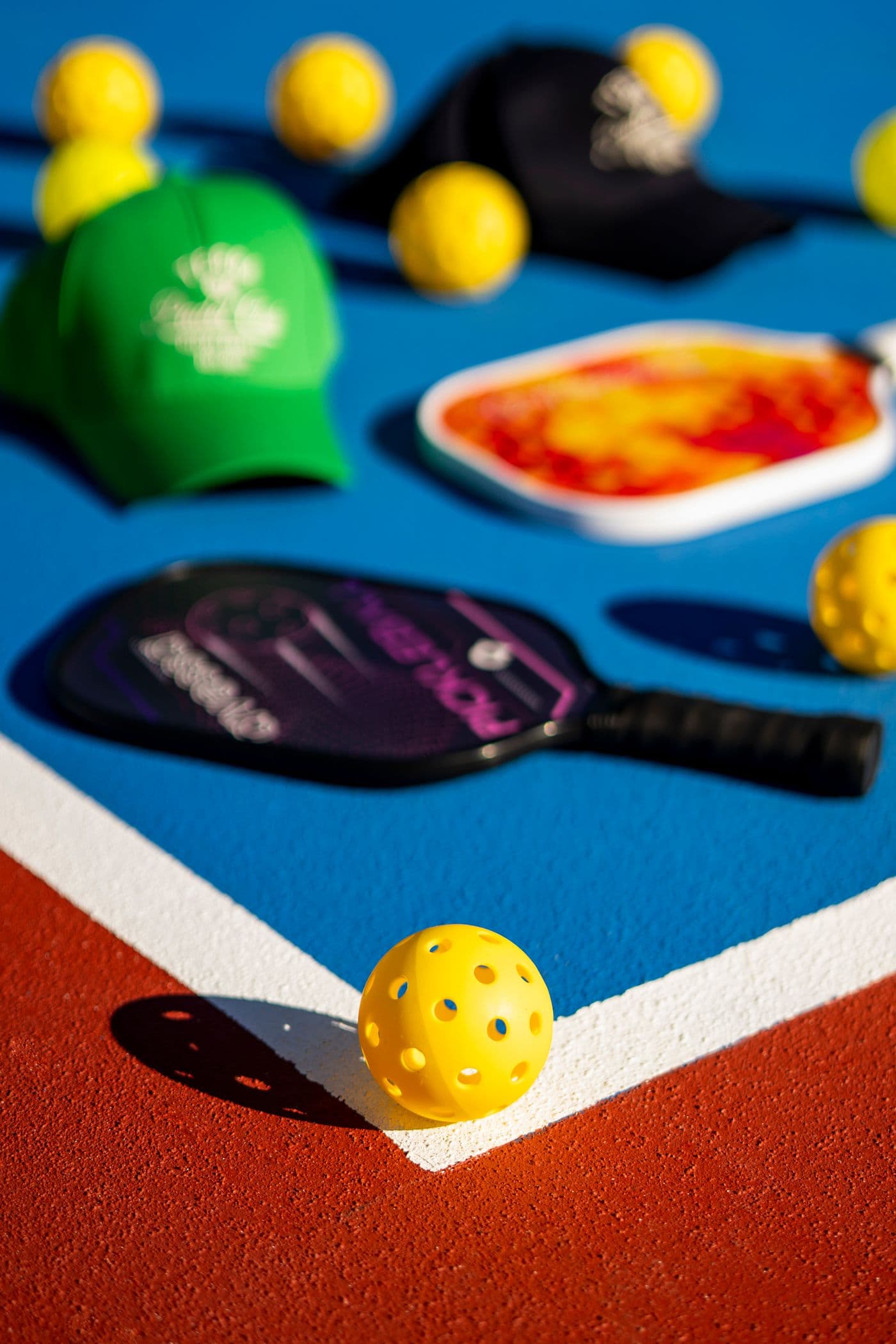 A yellow pickleball resting on the white line of a blue and red court, with paddles and caps behind it.