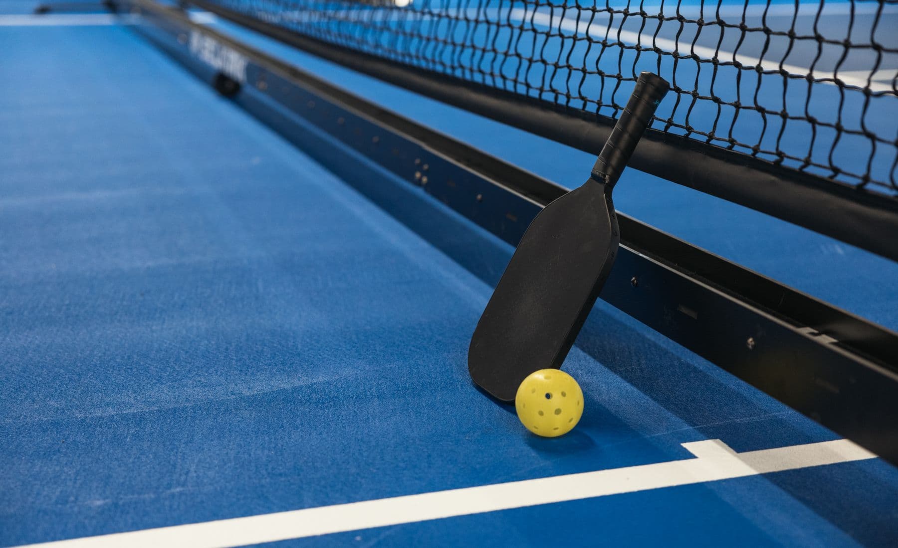 A paddle resting against a net post beside a pickleball on the court, no people in frame.